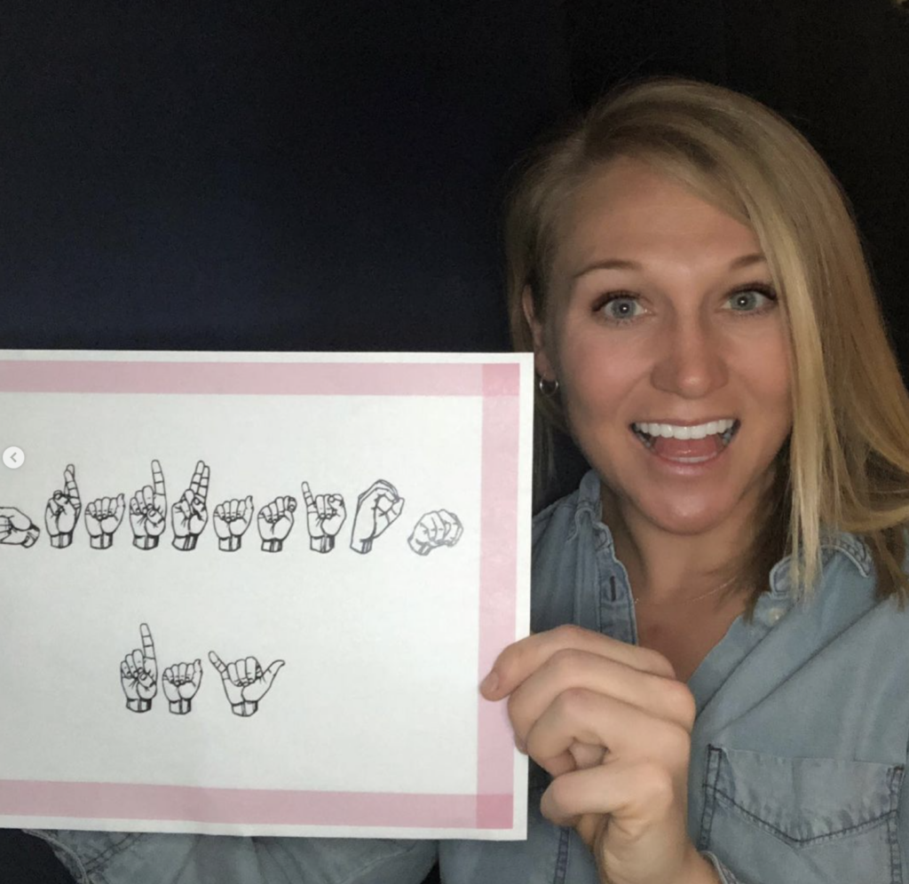 Blonde girl smiling holding a sign that has hands spelling out Graduation Day in ASL.